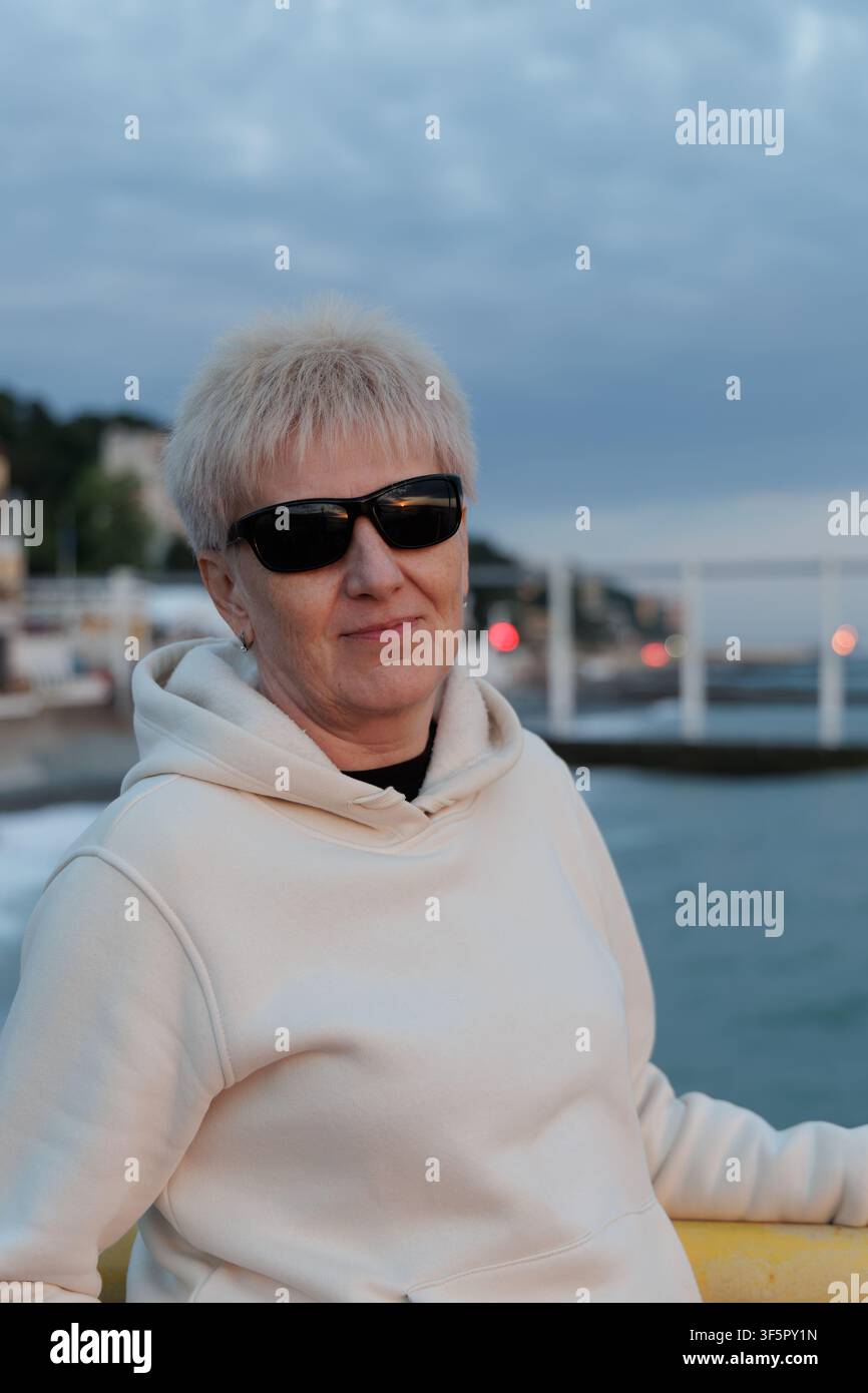 Sénior heureuse aux cheveux blonds courts portant un sweat à capuche décontracté et des lunettes de soleil, se relaxant sur une promenade côtière dans une station balnéaire européenne. Banque D'Images