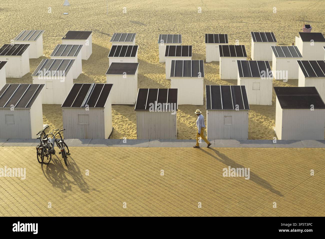 Des cabanes blanches aux toits sombres sont alignées en rangées ordonnées sur le sable d'Oostende, tandis qu'une personne marche le long du chemin pavé avec de longues ombres à côté de deux vélos garés Banque D'Images
