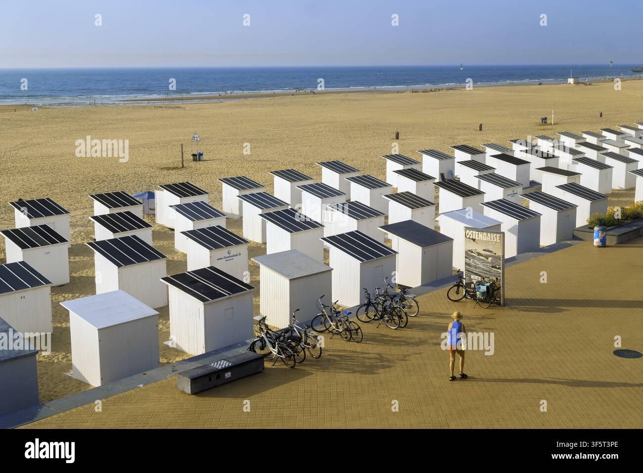 Des cabanes blanches aux toits sombres sont alignées en rangées ordonnées sur le sable d'Oostende, tandis qu'une personne marche le long du chemin pavé avec de longues ombres à côté de deux vélos garés Banque D'Images