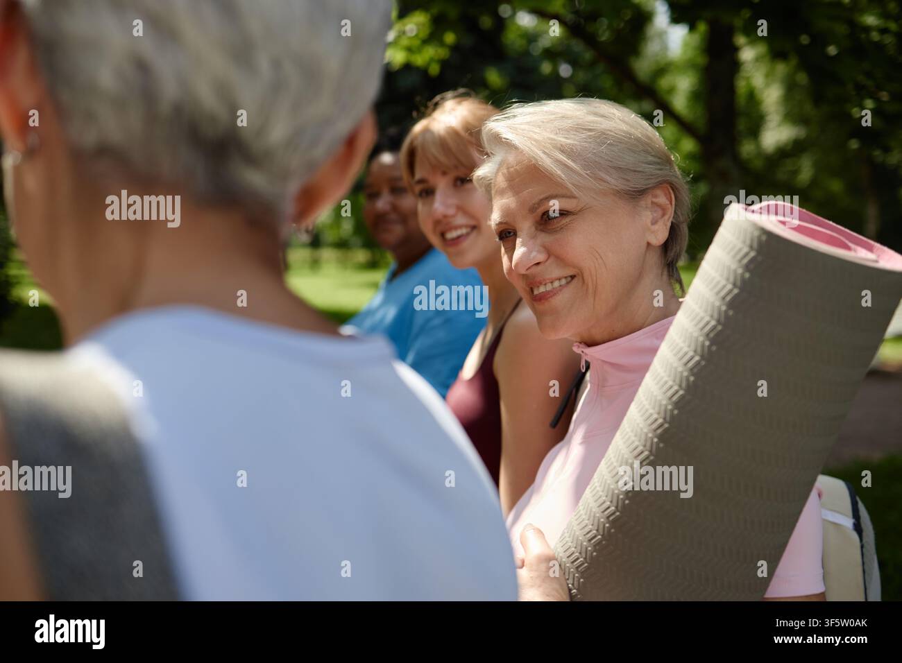 Sénior heureuse tenant un tapis de yoga et discutant avec des amis dans le parc Banque D'Images
