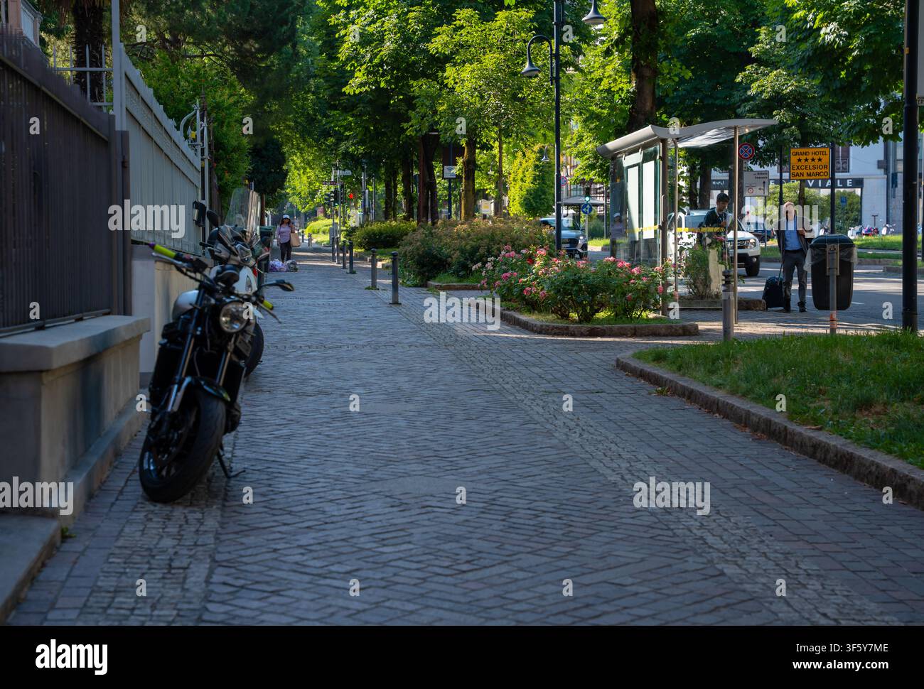 Bergamo, Italie – 16 juin 2026 : Scène de rue estivale lumineuse à Bergamo mettant en scène une moto garée, un trottoir pavé, des arbres verts luxuriants et des bus en verre Banque D'Images