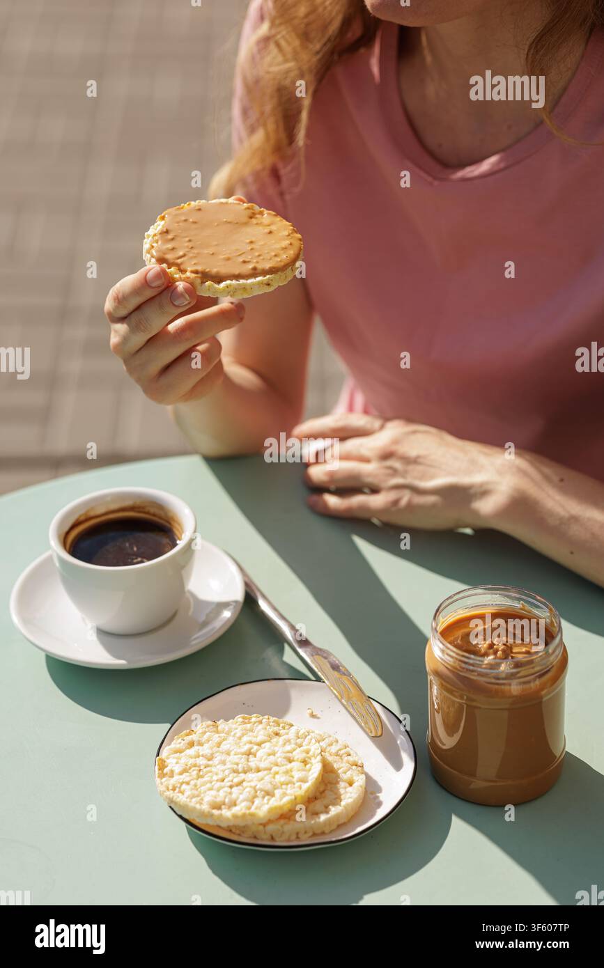Close-up d'une femme savourant un gâteau de riz au beurre de cacahuète et une tasse de café à une table extérieure, illustrant un en-cas sain simple et une pause café décontractée. Banque D'Images
