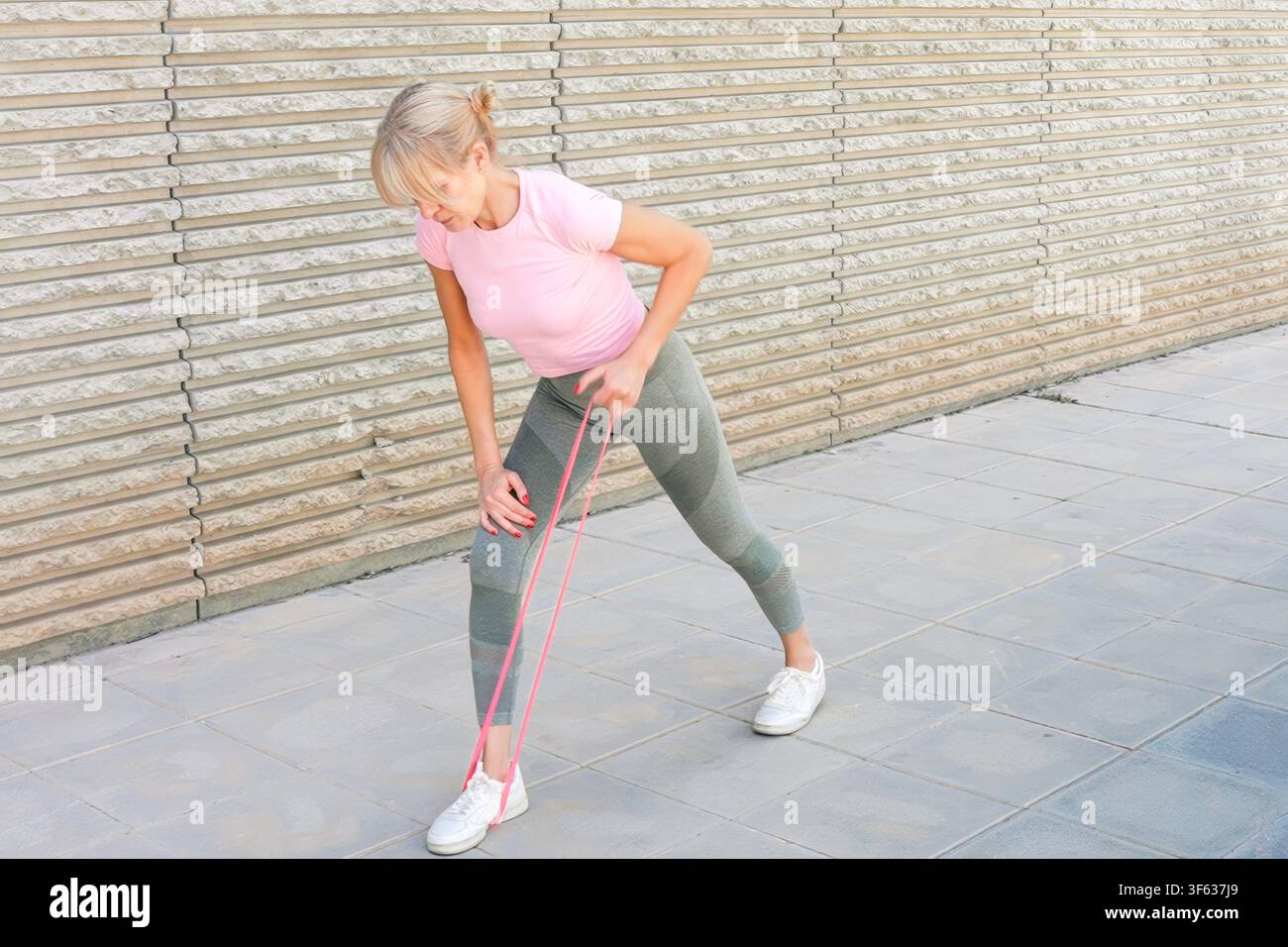 Une personne s'étirant avec des bandes de résistance dans un cadre urbain en plein air, concentrée et active, vue de côté, haut rose et leggings verts, pavé en béton Banque D'Images