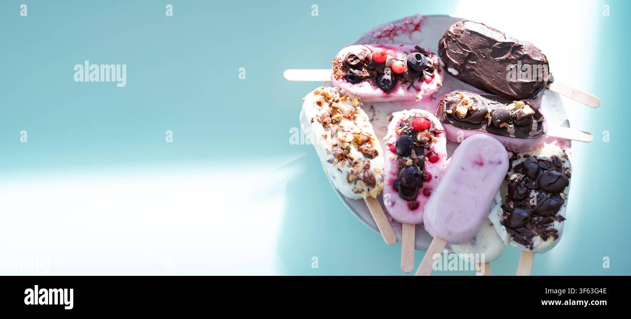 Ronde colorée de bâtonnets glacés maison au yaourt et au lait. Vanille et chocolat décorés de noix et de baies surgelées sur un fond bleu vif. Parfait pour une fête d'anniversaire d'enfants en été. Banque D'Images