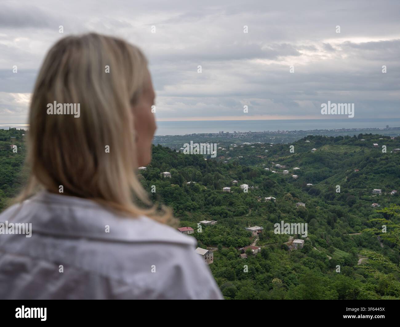 Femme blonde debout dos à la caméra, regardant les collines vertes luxuriantes couvertes de maisons dispersées, avec le ciel nuageux, les cités lointaines Banque D'Images