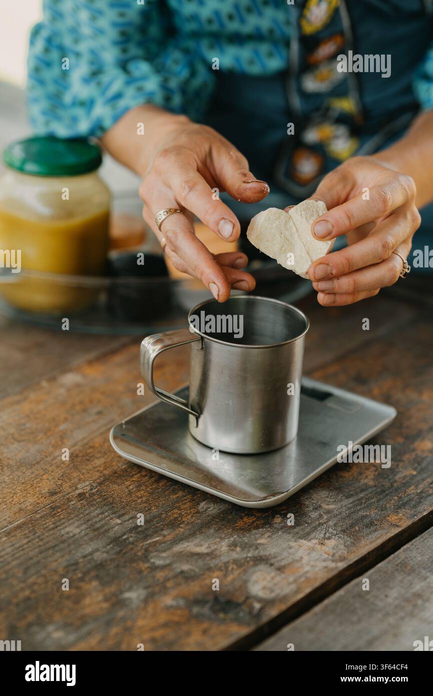Mains façonnant une pâte en forme de cœur au-dessus d'une tasse en métal sur une balance de cuisine avec une table rustique et une lumière chaude Banque D'Images