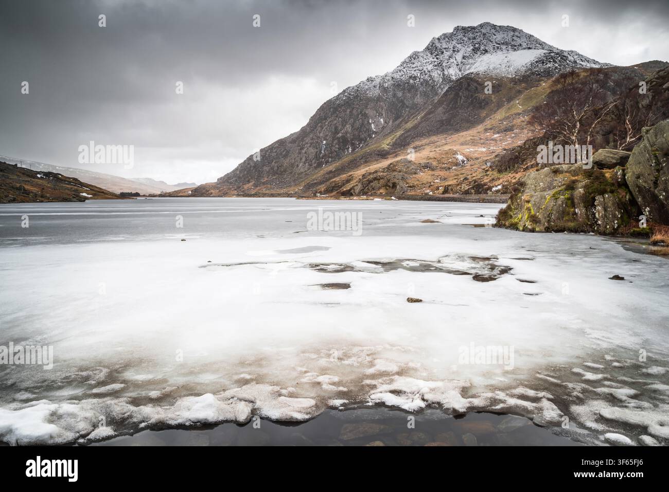 Vue hivernale dramatique sur la glace gelée de Llyn Ogwen en direction du sommet enneigé du mont Tryfan sous un ciel couvert. Temps hivernal Banque D'Images