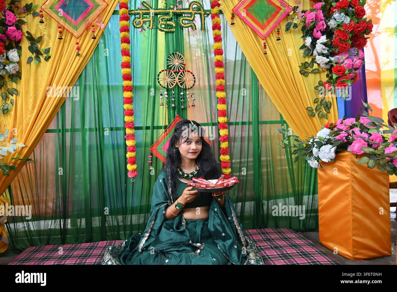 Jeune femme indienne en lehenga traditionnel vert assise pendant une cérémonie mehndi avec une assiette de henné, décorée de draperies jaunes et vertes et de motifs floraux indiens Banque D'Images