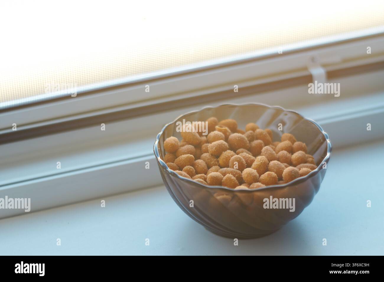 Vue rapprochée de cacahuètes grillées croustillantes dans un petit bol sombre posé sur un rebord de fenêtre lumineux avec une lumière naturelle filtrant à travers la vitre. Banque D'Images