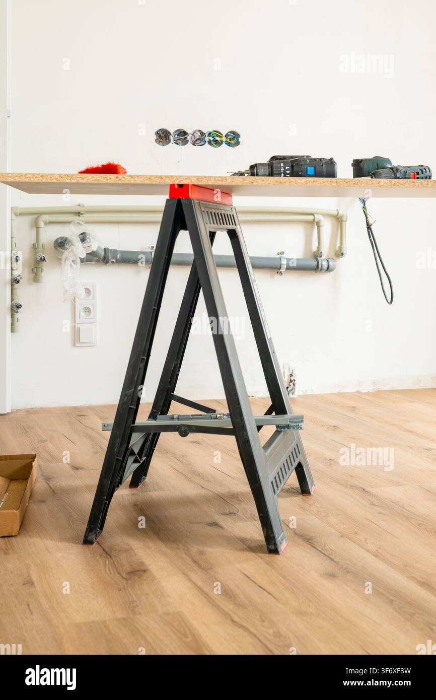 Construction d'une surface de travail temporaire à l'aide d'une échelle pliante noire et d'une planche de bois dans un cadre domestique. Les outils visibles incluent des perceuses et des composants électriques disposés sur le plan de travail. Banque D'Images