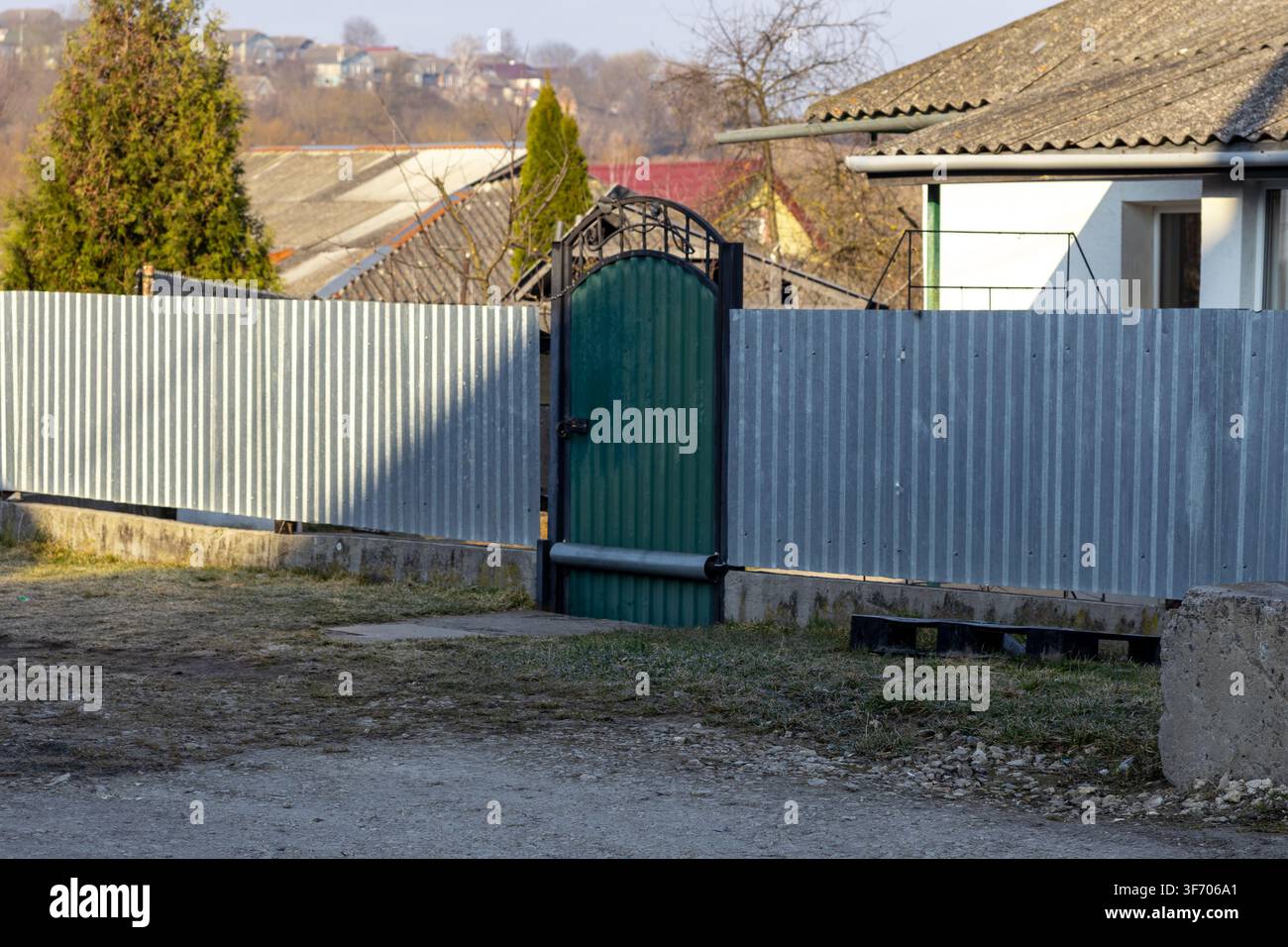 Panneaux de clôture en acier ondulé argenté avec une porte métallique verte dans un environnement de village rural, mettant en valeur les limites résidentielles, la sécurité domestique Banque D'Images