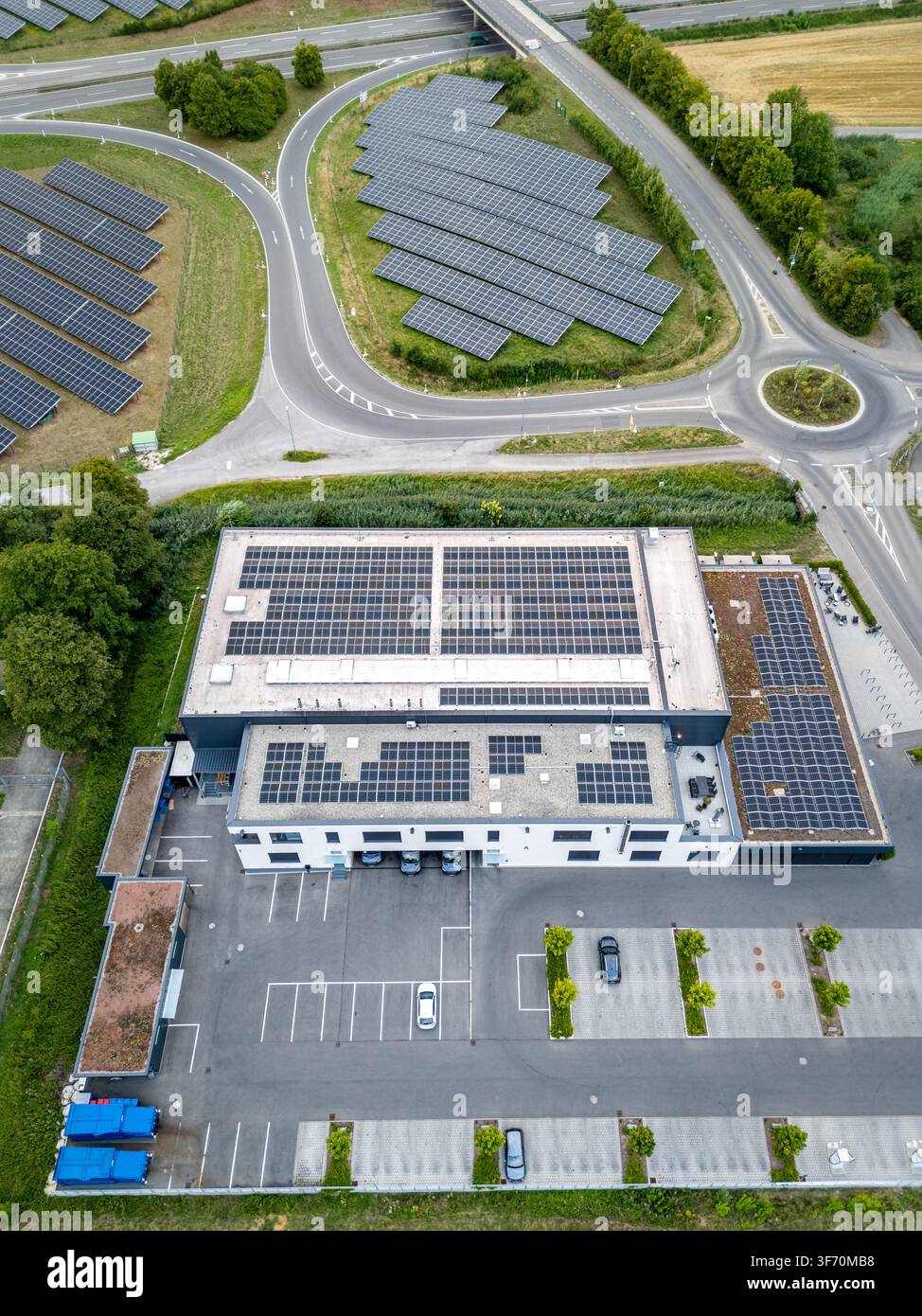Vue aérienne d'un grand toit de bâtiment commercial recouvert de panneaux photovoltaïques solaires, adjacent à une ferme solaire au sol, un parking et une route Banque D'Images
