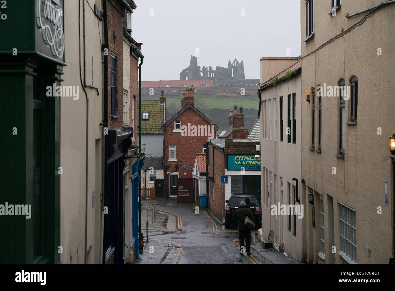 Un piéton solitaire navigue à travers une journée pluvieuse de décembre à Whitby, en Angleterre, encadré par les ruines historiques de l'abbaye de Whitby sur la colline au-dessus. Banque D'Images