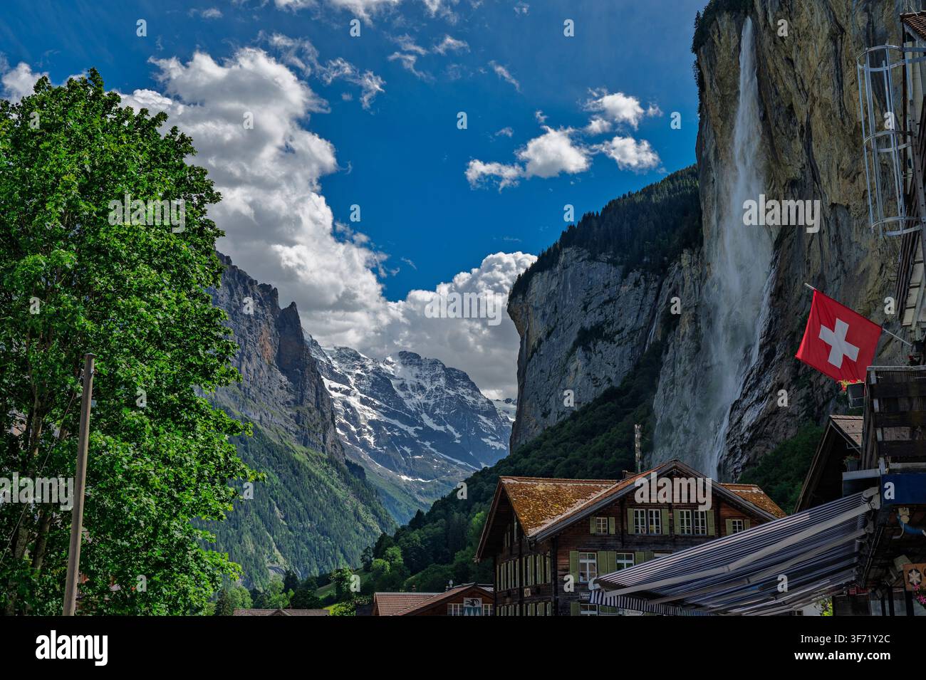 Une haute cascade dévale des falaises abruptes à côté de chalets en bois dans la vallée de Lauterbrunnen, avec un drapeau suisse flottant et des pics enneigés visibles au loin. Banque D'Images