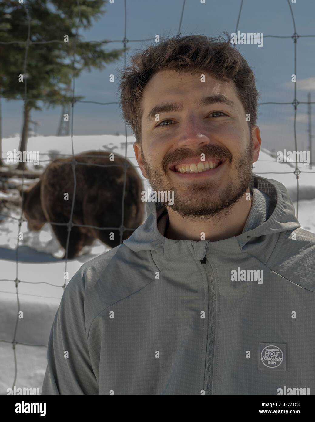 Vancouver, Canada – avril 2026 : Un visiteur pose pour une photo avec un ours grizzly broutant derrière une clôture en fil de fer au refuge faunique de Grouse Mountain. Banque D'Images