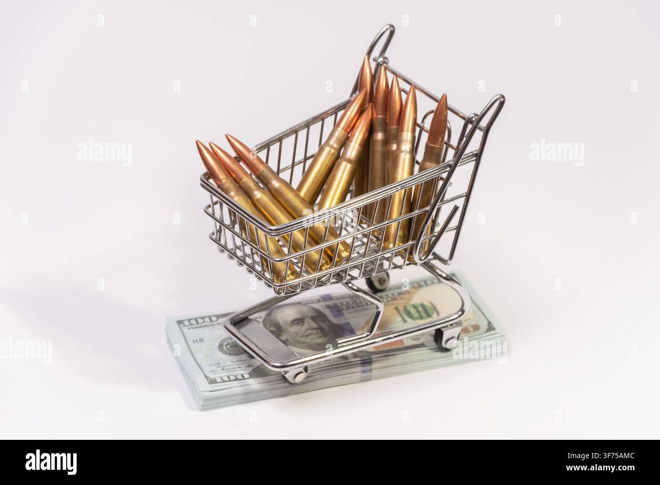 Un petit chariot de courses en métal chargé de cartouches de fusil en laiton est posé sur une pile de billets de cent dollars sur un fond blanc. Banque D'Images
