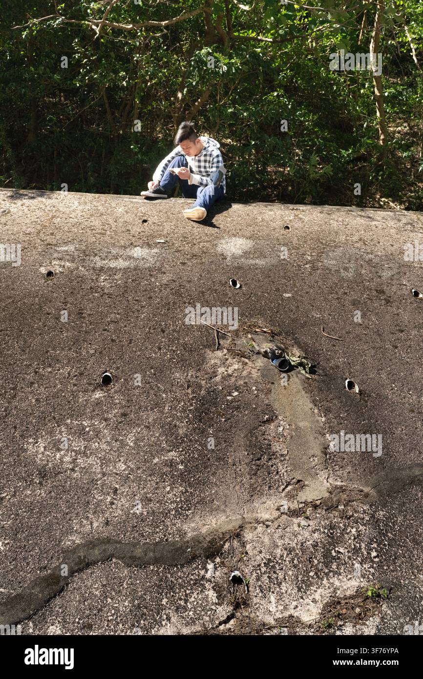 Un randonneur s'arrête pour vérifier son téléphone tout en étant assis sur un mur de protection en béton sur Temple Hill, à Kowloon, Hong Kong. Banque D'Images