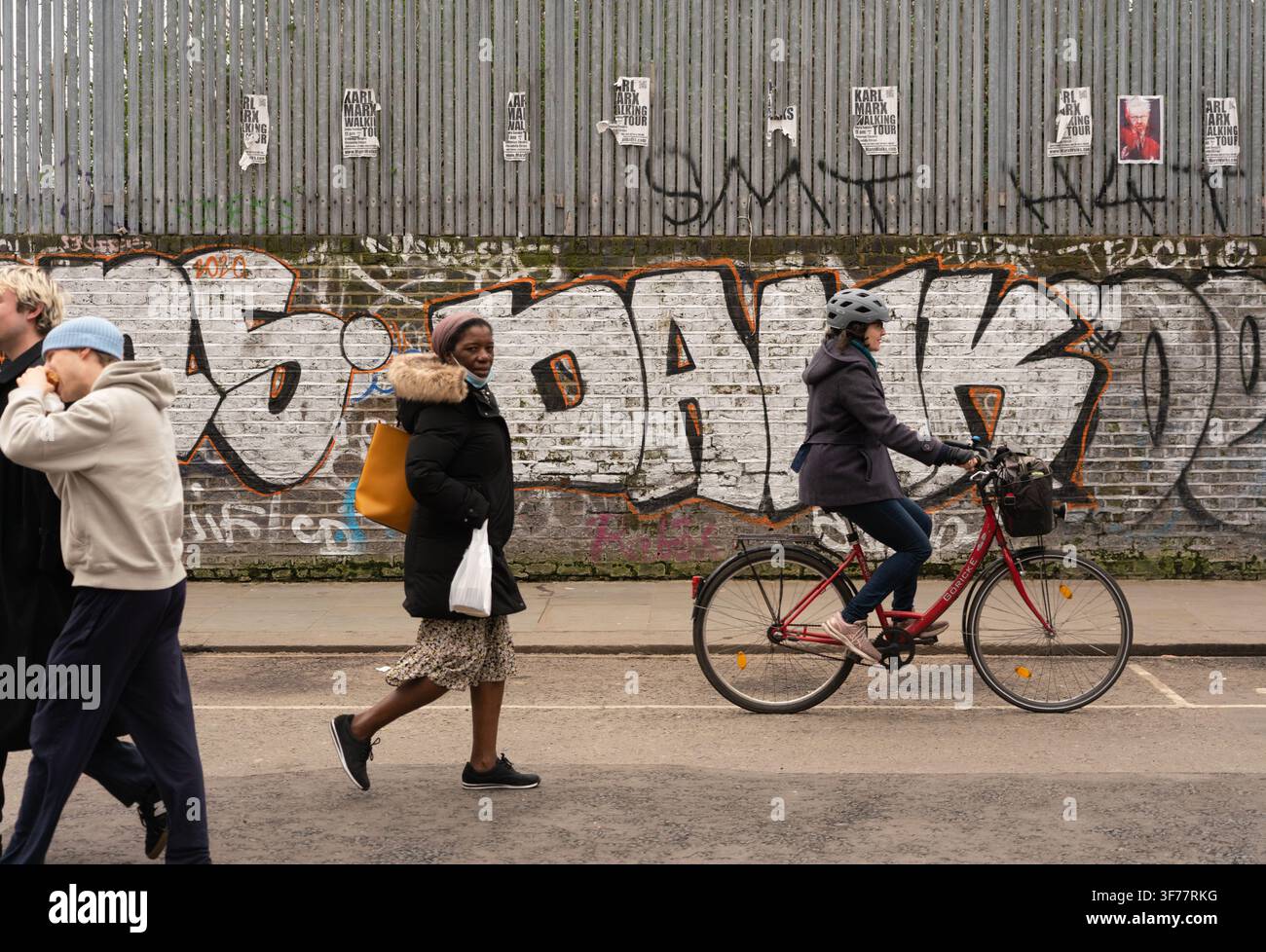 La vie urbaine quotidienne à Notting Hill : des navetteurs passent devant un mur couvert de graffitis, bordé d'affiches de la visite à pied Karl Marx sur Portobello Road. Banque D'Images