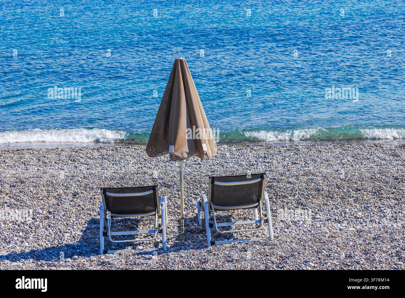 Belle vue sur deux transats et parasol fermé sur une plage de galets au bord de la mer. Crète. Grèce. Banque D'Images