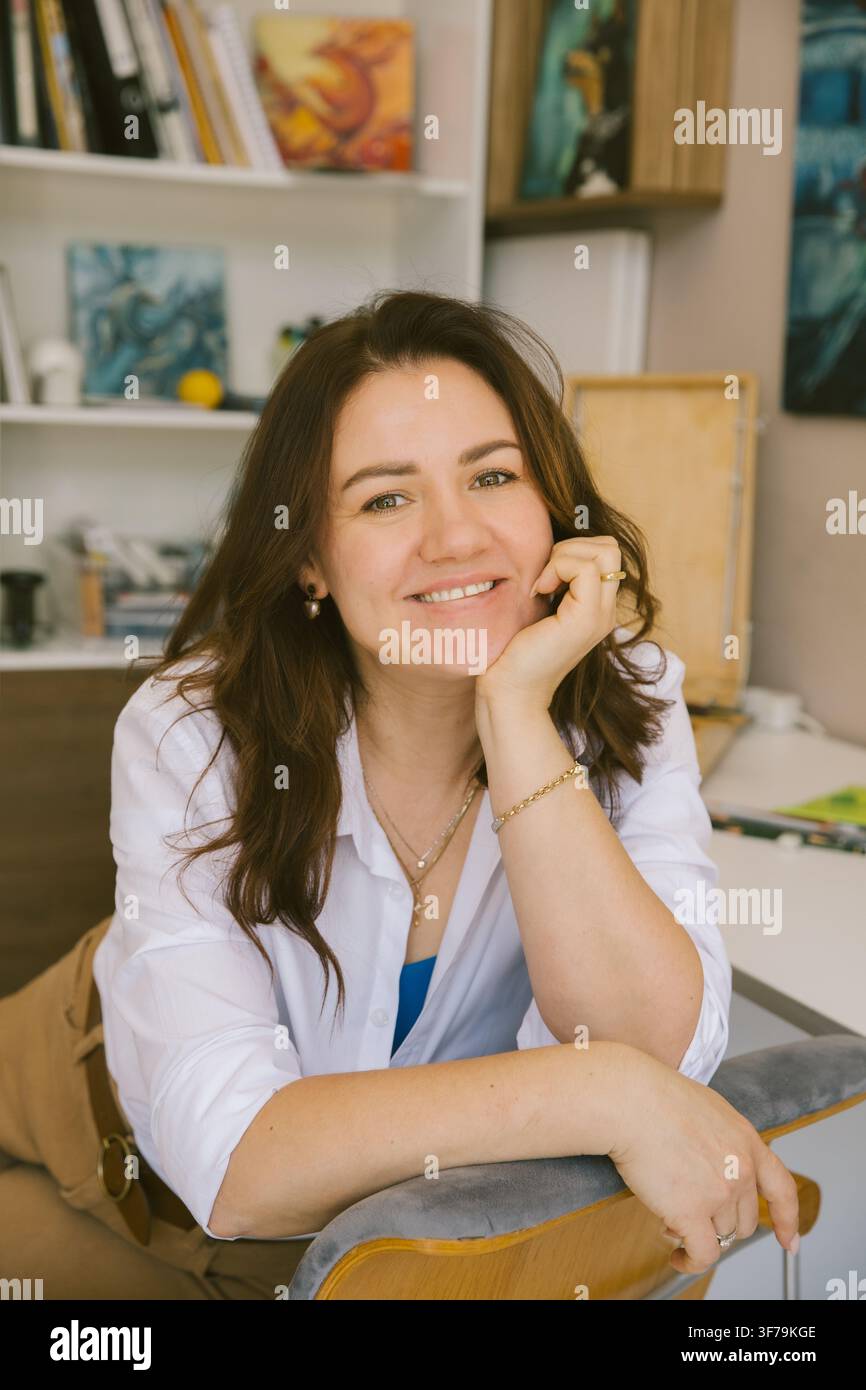 Portrait rapproché d'une artiste féminine souriante appuyée sur une chaise dans son studio créatif. Image de style de vie authentique d'une peintre contemporaine. Banque D'Images