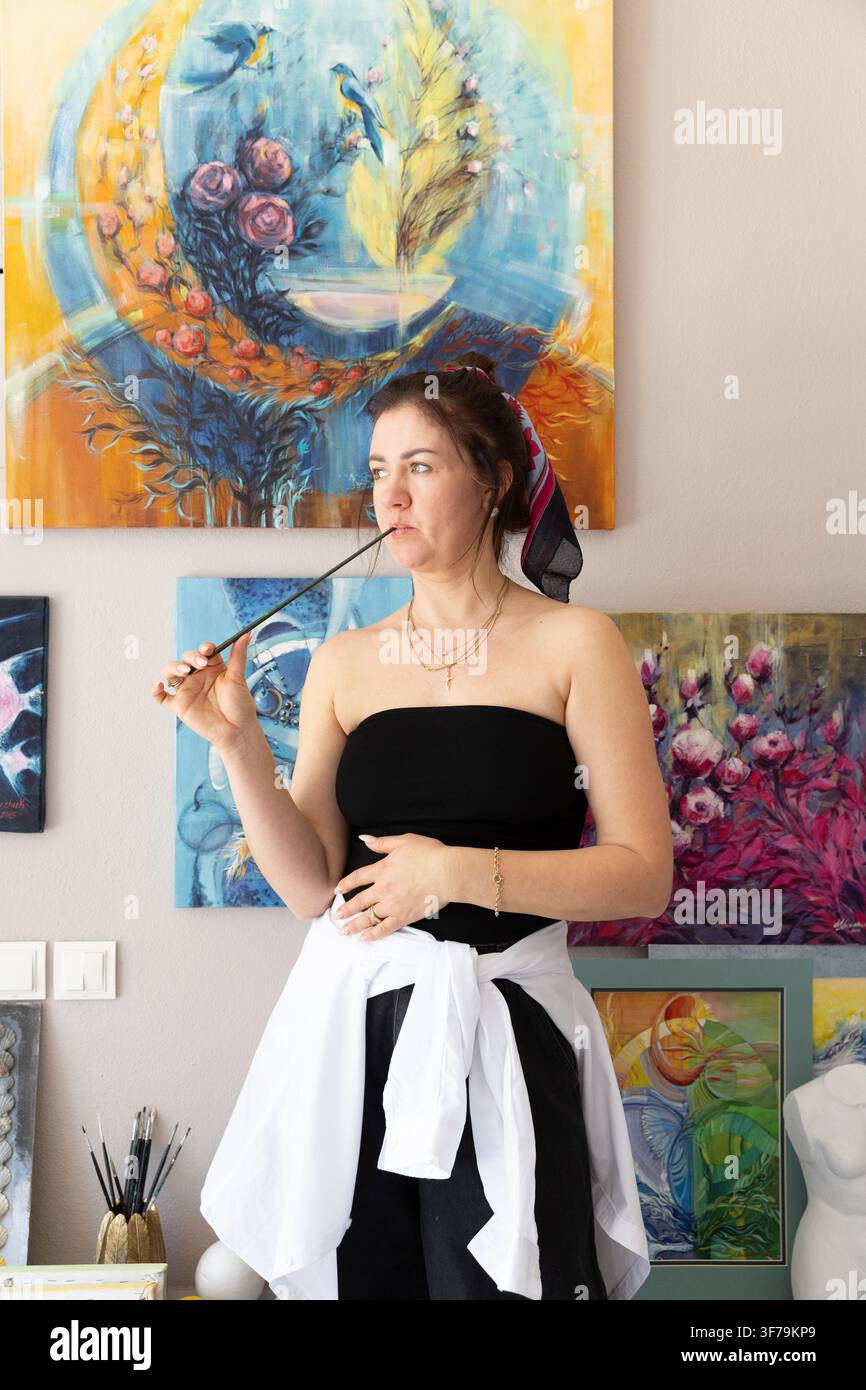 Artiste professionnelle debout dans son atelier créatif avec un pinceau, entourée de peintures et d'œuvres d'art originales colorées. Portrait authentique Banque D'Images
