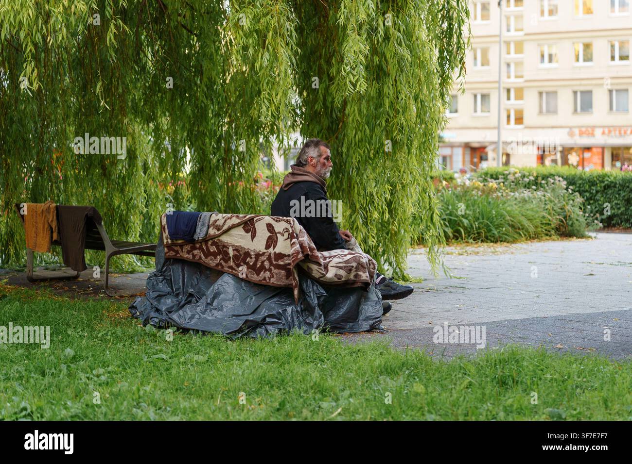Danszik, Pologne - 27 juillet 2026 : Sans-abri reposant sur un banc de parc urbain, enveloppé dans des couvertures sous un arbre, représentant la lutte sociale et la pauvreté urbaine, survie Banque D'Images