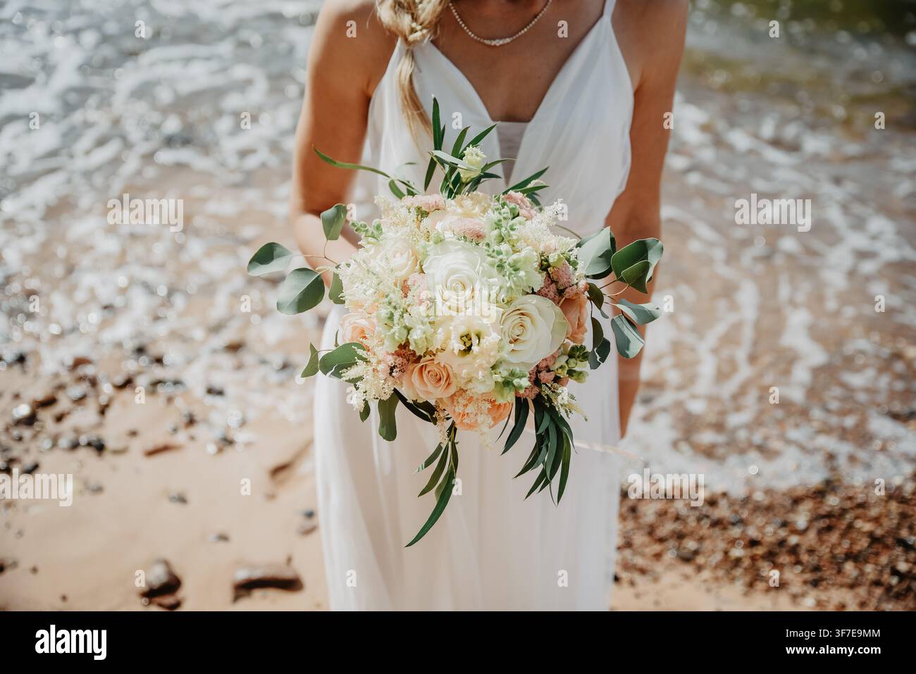 Mariée élégante tenant un bouquet de mariage pastel romantique sur une plage ensoleillée, combinant paysage côtier, mode nuptiale et esthétique de mariage de destination Banque D'Images