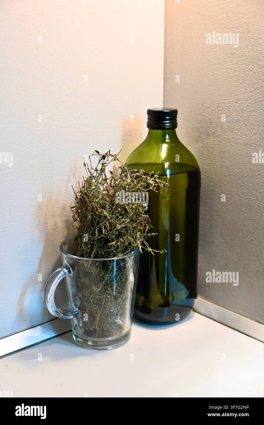 Une photographie verticale capturant une composition de nature morte mettant en scène une tasse en verre transparent remplie d'herbes sèches et une haute bouteille en verre vert avec un bouchon noir, placées dans un coin contre des murs de couleur claire. Banque D'Images
