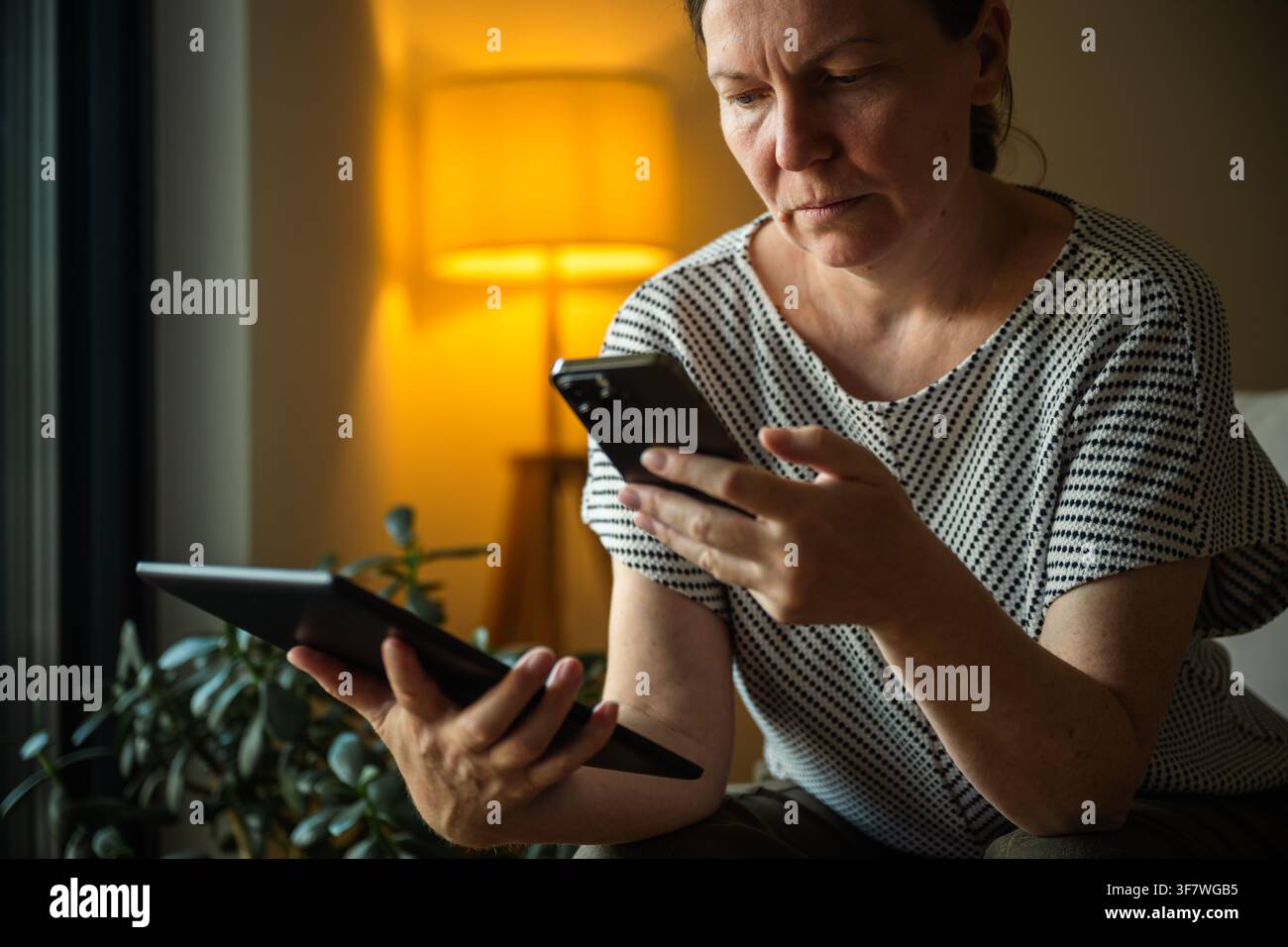 Femme mûre utilisant une tablette numérique et un smartphone pour synchroniser les données entre les appareils à la maison. Connectivité inter-appareils, technologie cloud et digital Banque D'Images