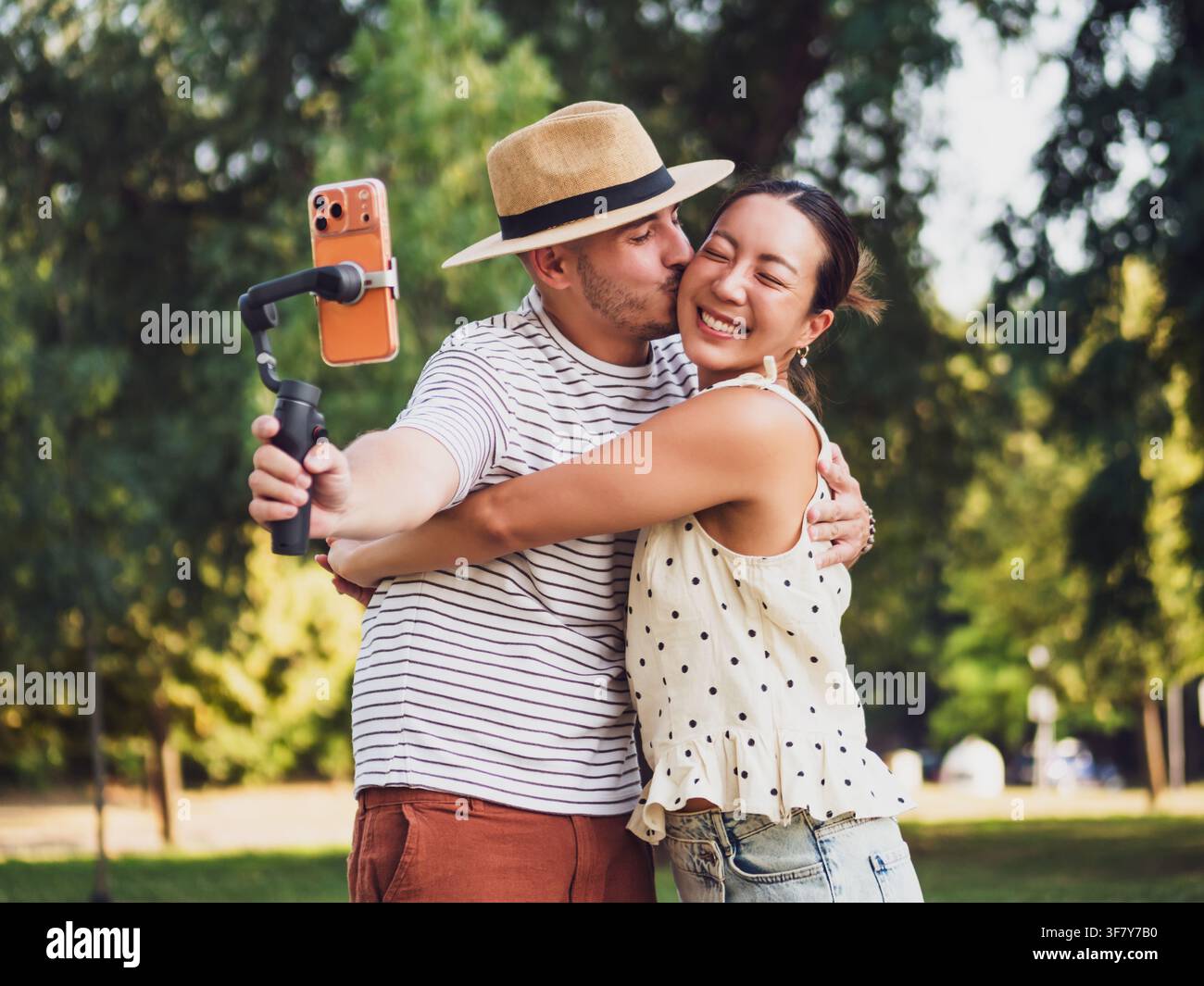 Jeun couple joyeux s'embrassant et riant tout en se filmant avec un stabilisateur de smartphone en plein air. Banque D'Images