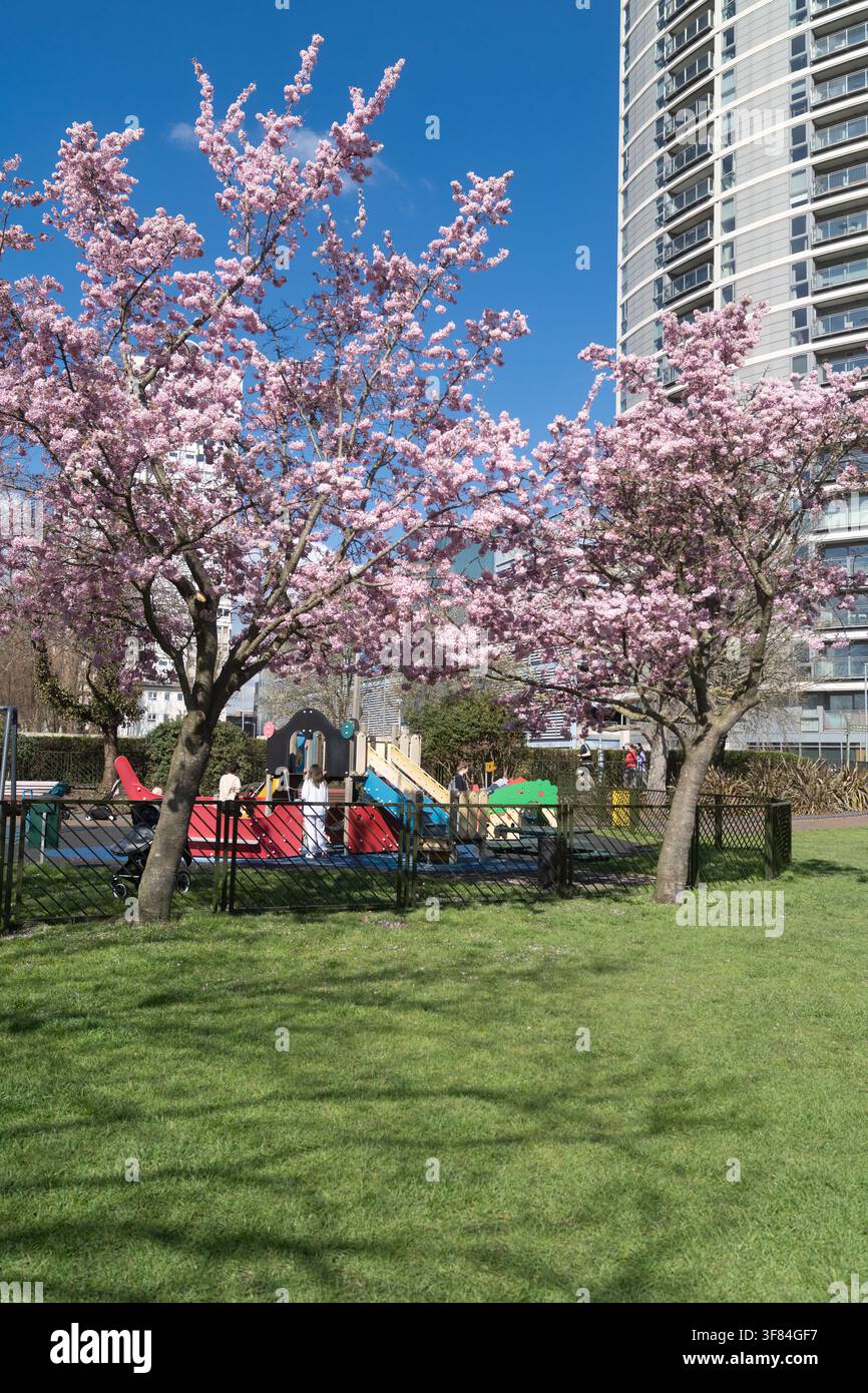 Les familles profitent du soleil printanier sur le terrain de jeu du King George’s Park à Wandsworth, encadré par des fleurs de cerisier et la skyline de la Ram Brewery. Banque D'Images