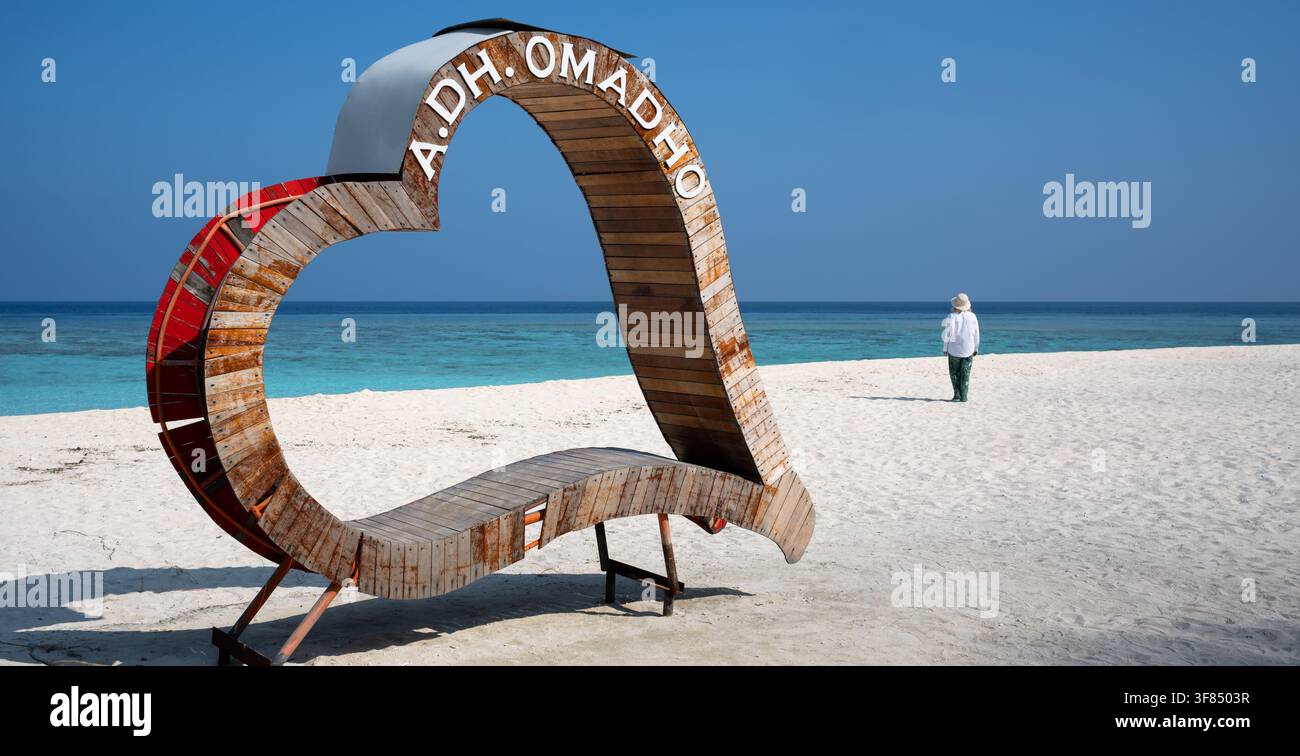 Une grande structure en bois en forme de cœur se dresse sur une plage de sable blanc, arborant le texte A.DH. OMADHO peint en blanc le long de sa courbe supérieure. Dans la ba Banque D'Images