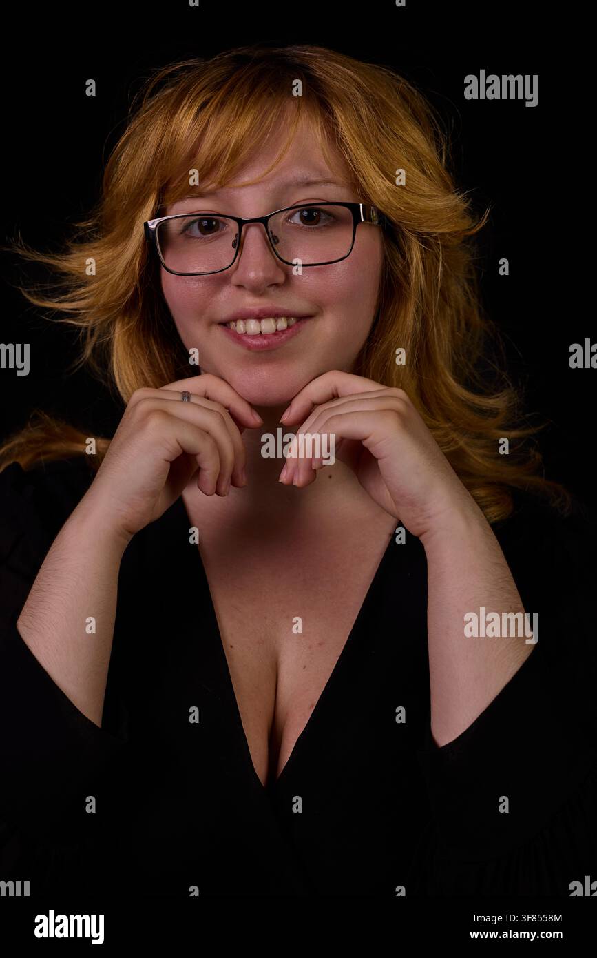 Jeune femme aux cheveux blonds souriant et reposant son menton sur ses mains contre un fond noir Banque D'Images