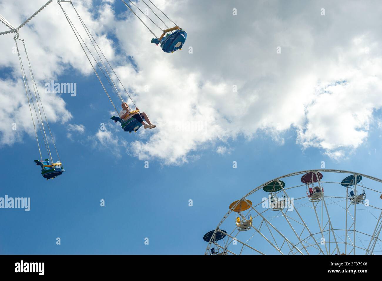 Une fille sur une balançoire au-dessus d'une grande roue contre un ciel bleu dans un parc de la ville. Banque D'Images