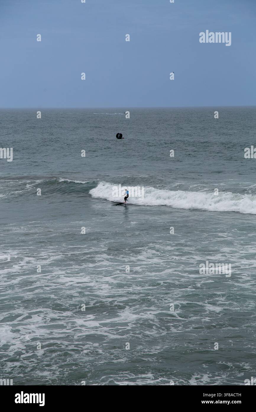 Surfeur chevauchant une petite vague de l'océan sous un ciel nuageux, entouré de mer agitée et d'eau mousseuse. Le surfeur distant est capturé face à l'océan Banque D'Images