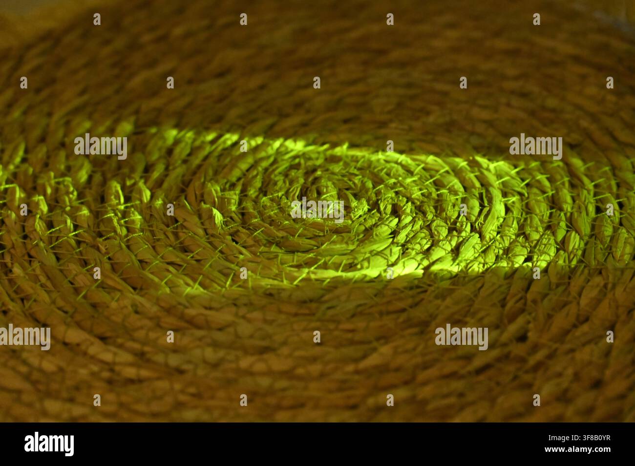 Photographie macro rapprochée commerciale d’un rayon de lumière vert vibrant ou laser abstrait traversant la texture complexe et tourbillonnante d’un set de table ou d’un dessous de verre rond en paille tissée, sur un fond sombre. Banque D'Images