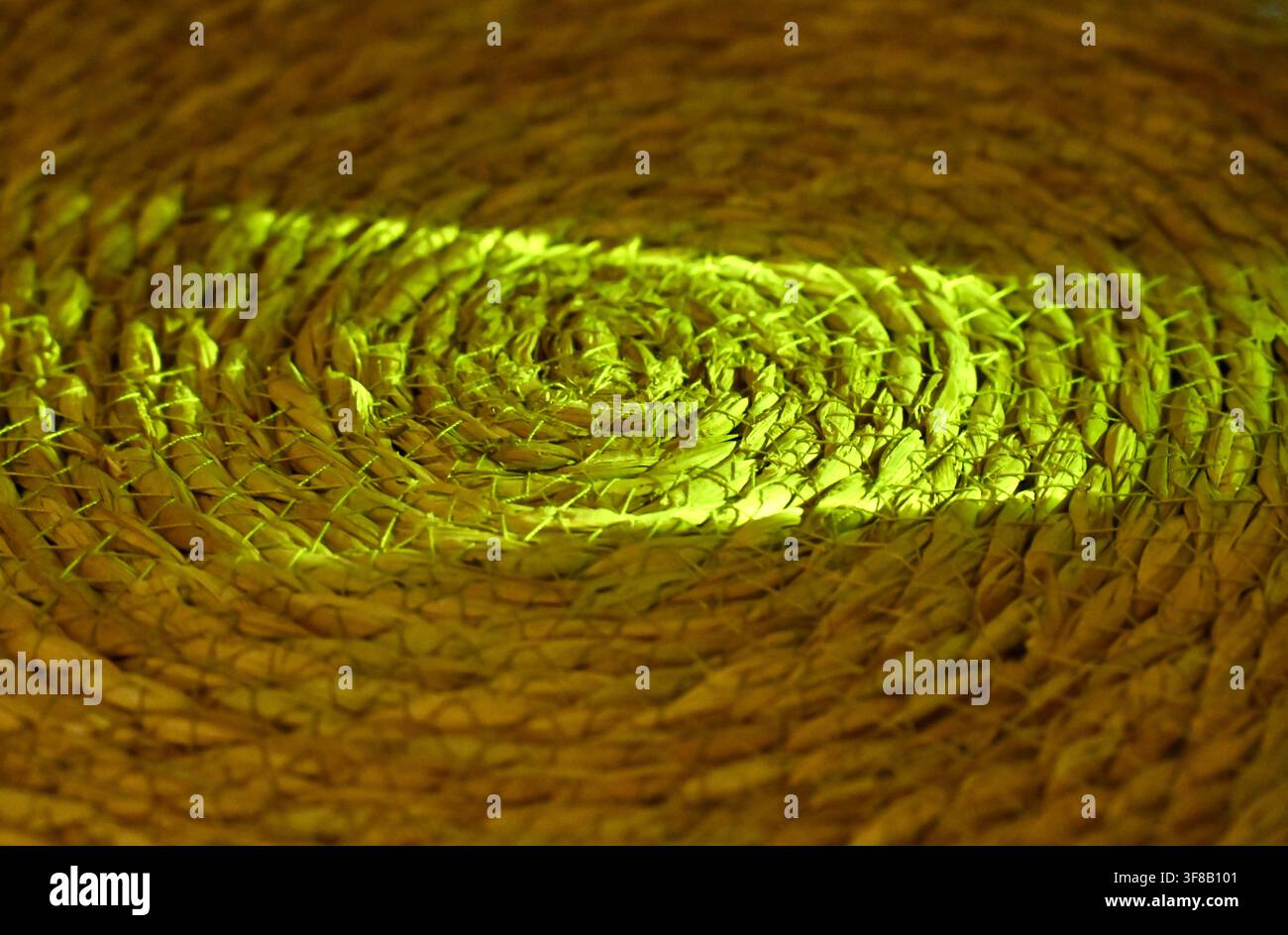 Photographie macro rapprochée commerciale d’un rayon de lumière vert vibrant ou laser abstrait traversant la texture complexe et tourbillonnante d’un set de table ou d’un dessous de verre rond en paille tissée, sur un fond sombre. Banque D'Images