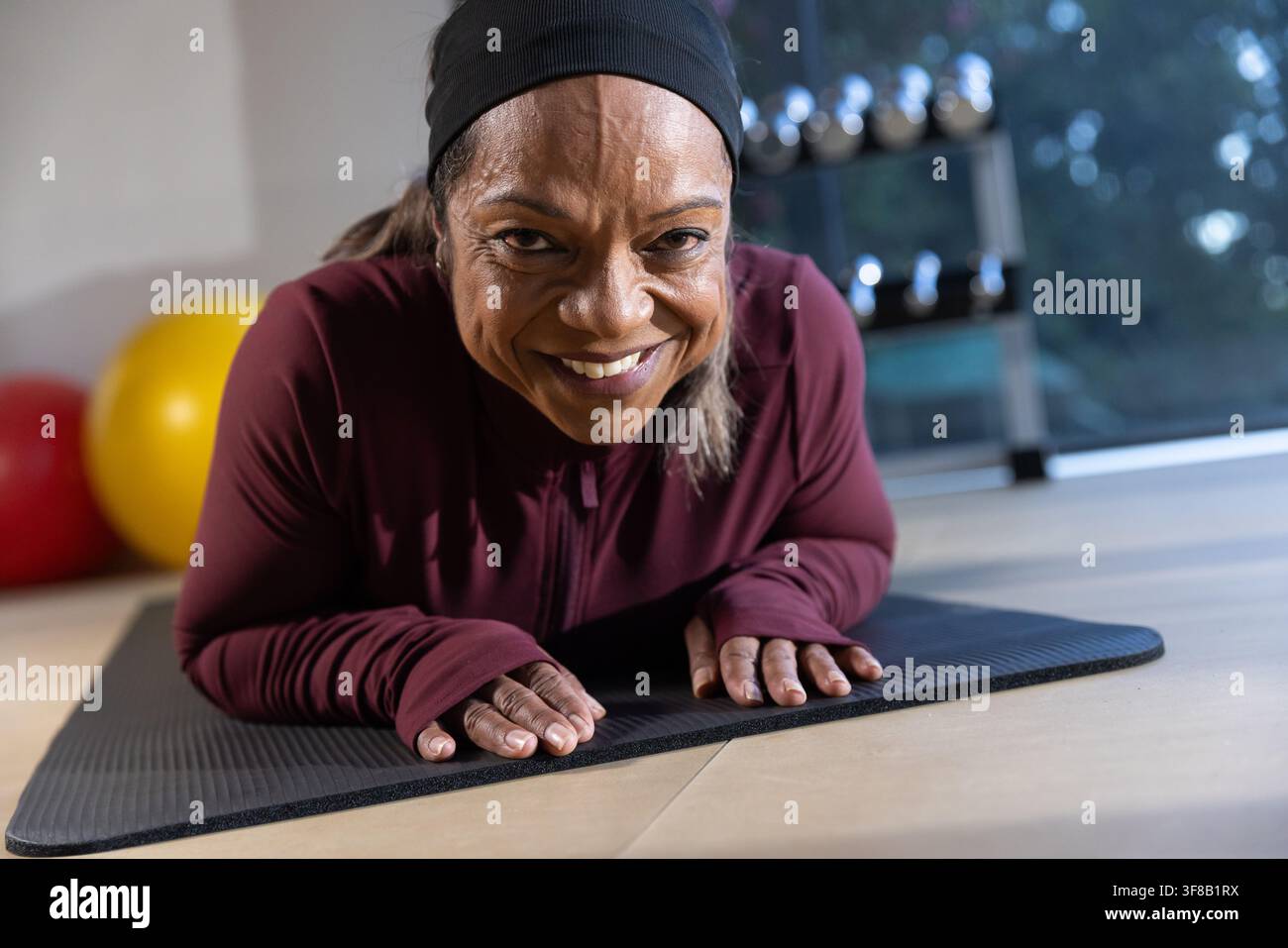Femme mûre effectuant des planches sur les avant-bras sur un tapis noir dans une salle de sport avec des haltères, portant un haut bordeaux. Fitness, studio, abdominals, stabilité, poids, naturel, lig Banque D'Images