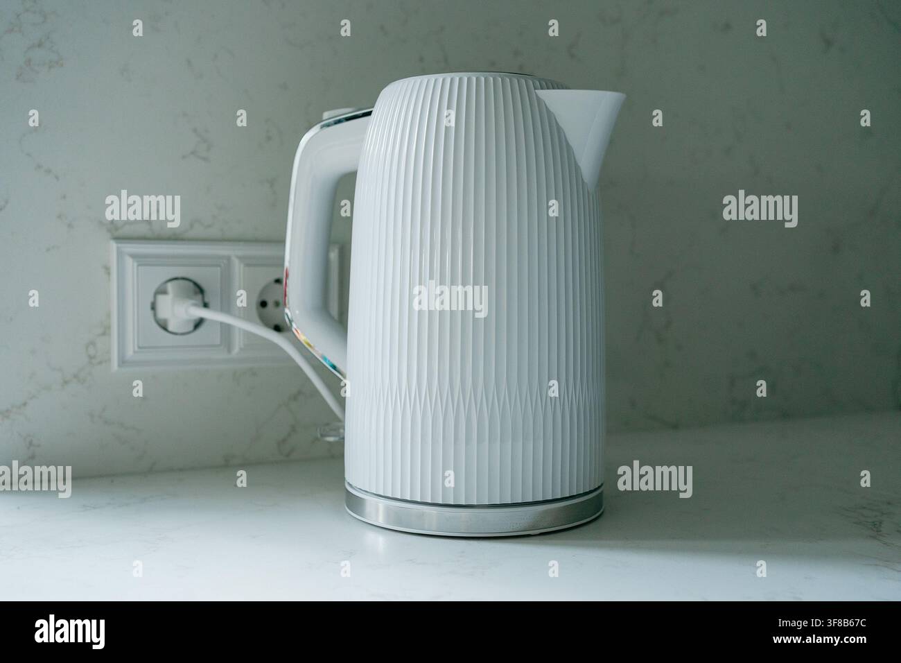 Théière électrique blanche posée sur un plan de travail connectée à une prise de courant dans un intérieur moderne. Image parfaite pour un magasin d'électroménager, cuisine Banque D'Images