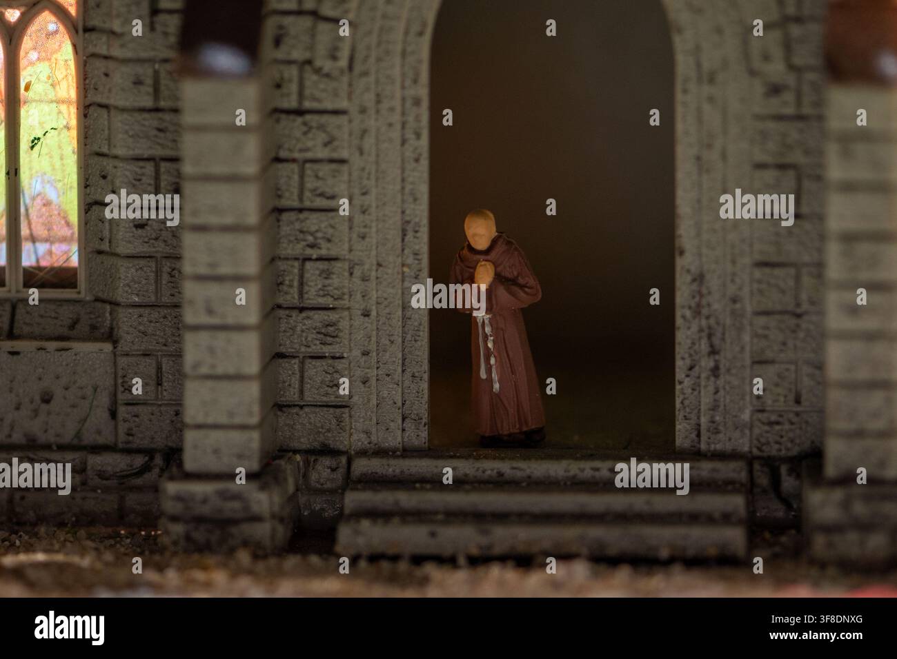 Une petite figurine de moine aux mains jointes se tient dans l'entrée sombre d'un modèle d'église en pierre à côté d'une vitre en vitrail lumineuse. Banque D'Images