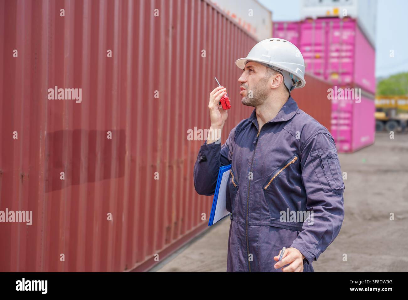 Ouvrier d'entrepôt communiquant par talkie-walkie dans une cour d'expédition. Idéal pour les thèmes de logistique, d'opérations et de gestion industrielle dans les affaires et Banque D'Images