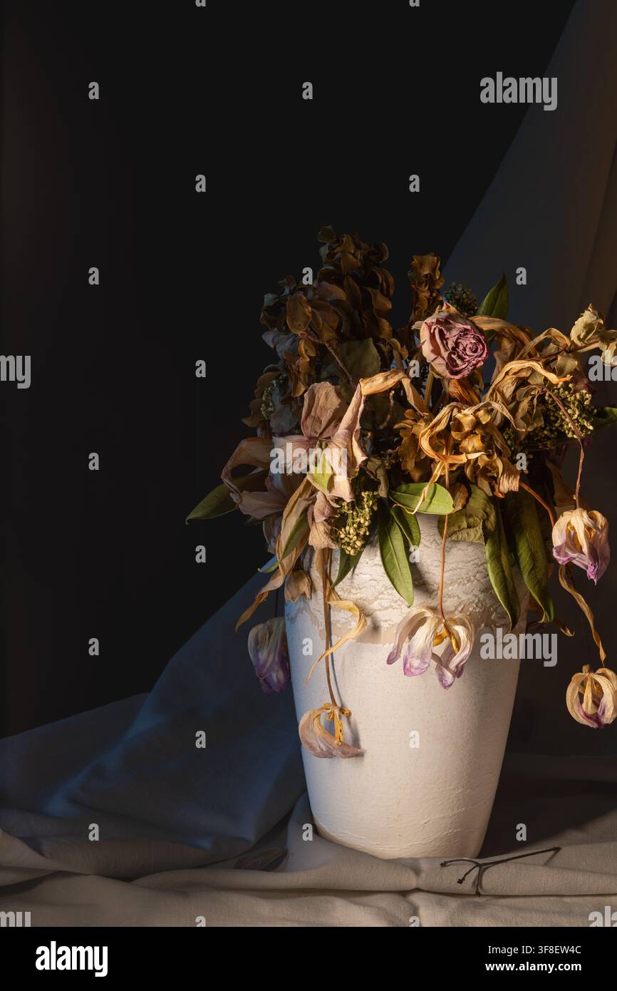 Vase blanche avec des fleurs séchées à côté d’un rideau en tissu bleu-vert et d’une nappe blanche, fond sombre Banque D'Images