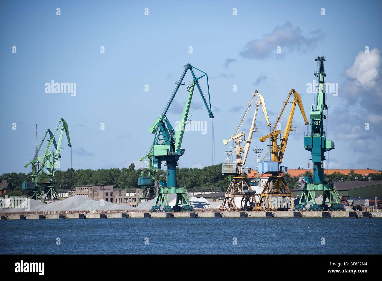 De grandes grues portuaires bordent le front de mer alors qu'elles chargent et déchargent des marchandises en vrac dans un port industriel actif, avec des tas de granulats, un ciel bleu et une eau calme Banque D'Images