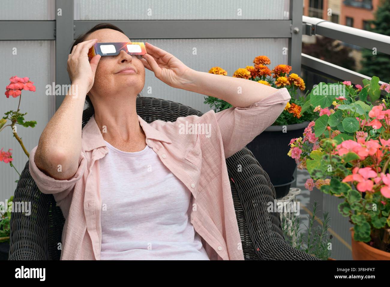 Femme d'âge moyen assise sur une chaise sur le balcon, portant des lunettes d'éclipse solaire, souriant et regardant vers le ciel entourée de plantes en pot Banque D'Images