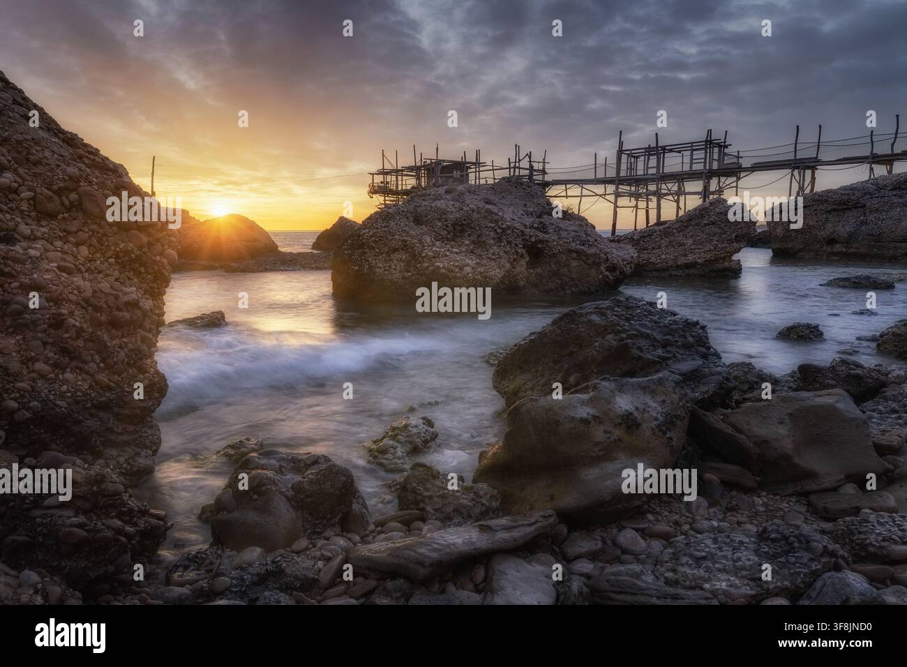 Vue du Trabocco Spezzacatena au coucher du soleil avec une côte rocheuse, des vagues et une lumière dorée à Rocca San Giovanni, province de Chieti, Italie. Banque D'Images