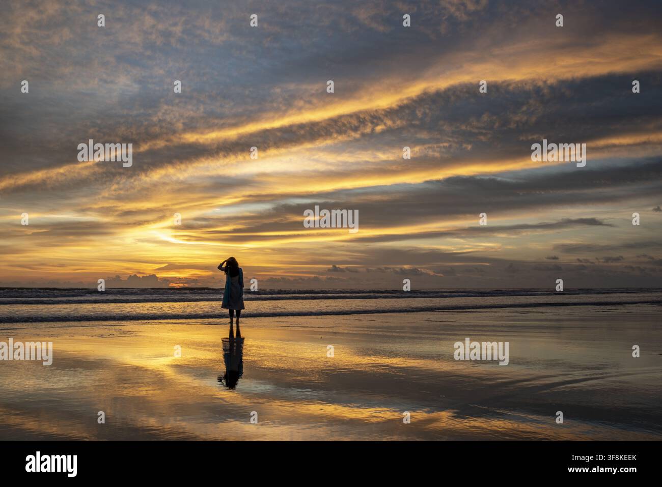 Vue d'une femme debout sur le sable mouillé d'Inani Beach au coucher du soleil, avec une lumière dorée se reflétant sur l'eau et un ciel dramatique dans le district de Cox's Bazar Banque D'Images