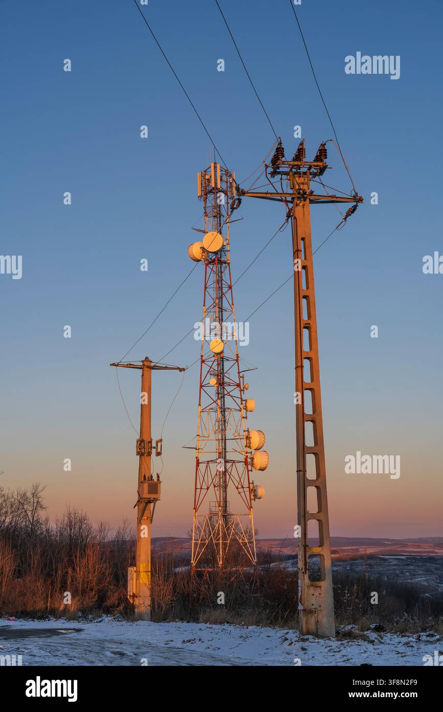 Une station de base cellulaire et des poteaux électriques haute tension se dressent contre un coucher de soleil d'hiver. Cette image représente la convergence technologique, une connexion mondiale Banque D'Images