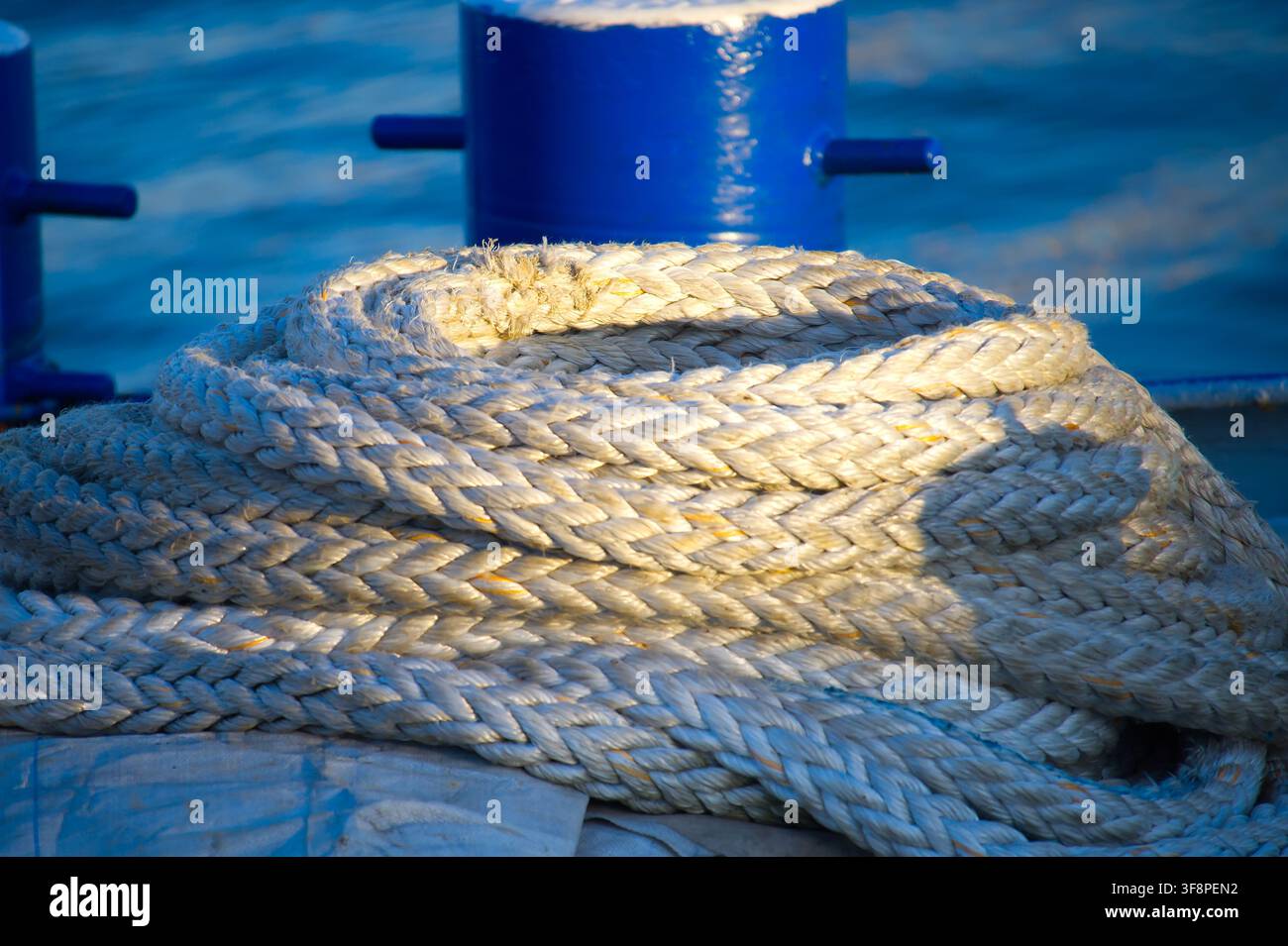 Corde d'amarrage épaisse enroulée reposant sur le pont d'un bateau à côté d'une bitte bleue, éclairée par une lumière chaude du soleil avec la mer en arrière-plan. Détail nautique en gros plan Banque D'Images