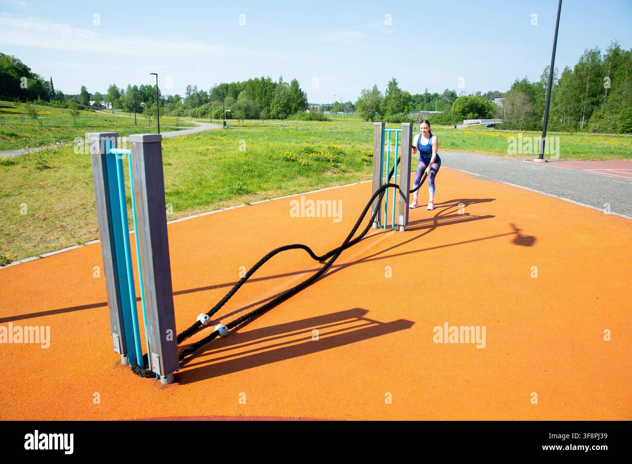 Entraînement de femme avec des cordes de bataille sur le complexe sportif extérieur orange avec terrain de basket-ball. Vue grand angle du parc de fitness urbain par jour ensoleillé. Banque D'Images