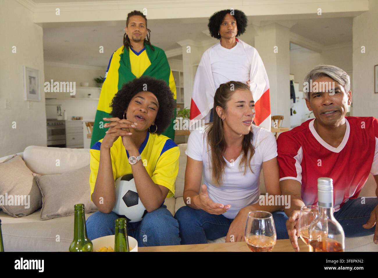 Des amis divers portant des maillots regardant un match de football à la maison, tenant un ballon de football et des bouteilles de bière Banque D'Images